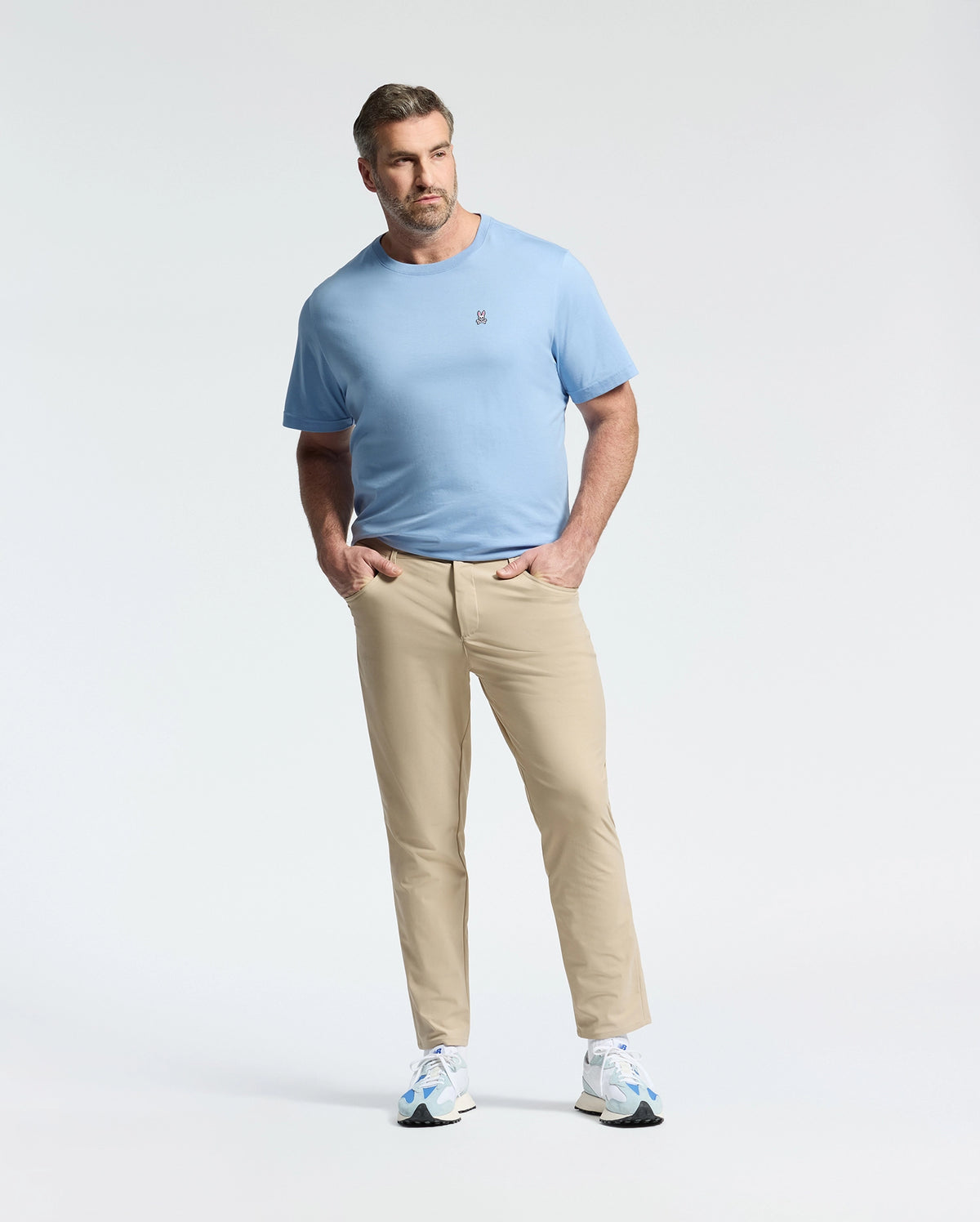 Wearing a Psycho Bunny BIG AND TALL CLASSIC CREW NECK TEE in light blue and beige pants, a man stands confidently with hands in pockets against a white background. He sports white sneakers, short hair, and a beard, showcasing effortless style.