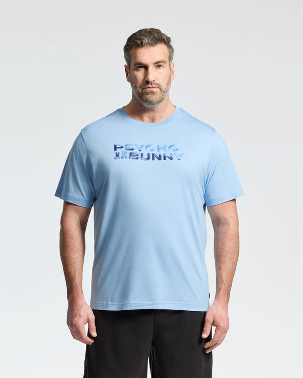 A man with light brown hair and a beard stands against a plain light background, wearing Psycho Bunny’s MEN'S BIG AND TALL BERNI EMBROIDERED WORDMARK GRAPHIC TEE (B9U417F200) in light blue, paired with black pants.
