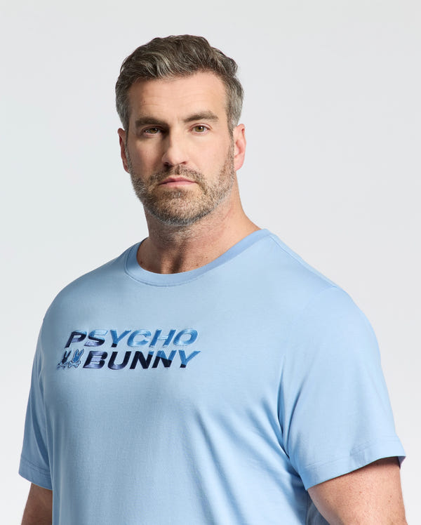 A man with short brown hair and a beard wears the Psycho Bunny Mens Big and Tall Berni Embroidered Wordmark Graphic Tee (B9U417F200), looking directly at the camera with a neutral expression against a plain, light background.