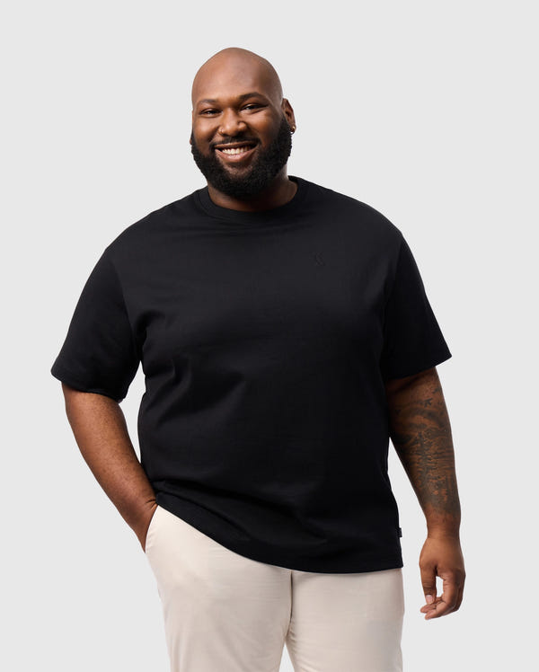 A person with a beard smiles against a plain background, wearing Psycho Bunny's BIG AND TALL DUKE OVERSIZED TEE in heavyweight Pima cotton and light-colored pants. One hand is in their pocket, and an embroidered chest bunny brings a playful touch to their look.