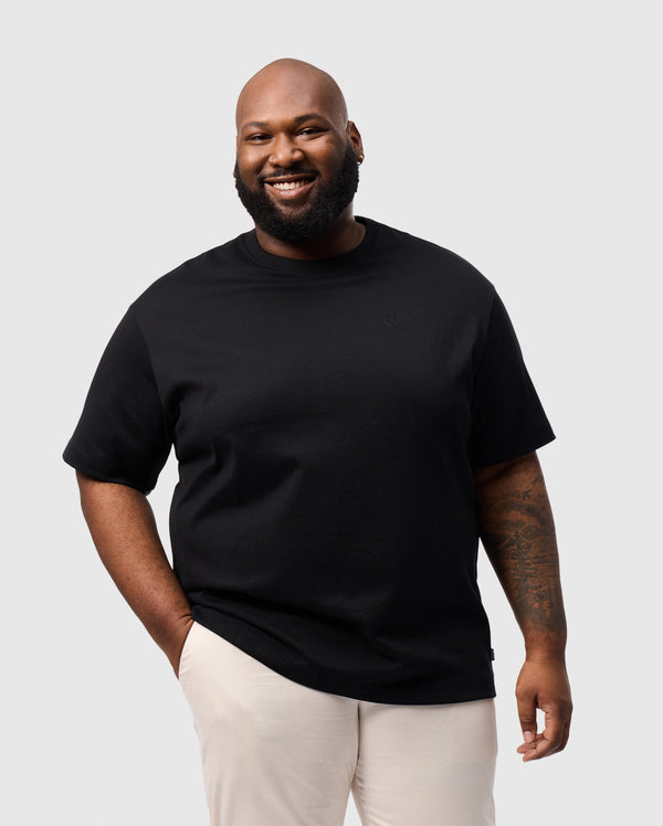 A person with a beard smiles against a plain background, wearing Psycho Bunny's BIG AND TALL DUKE OVERSIZED TEE in heavyweight Pima cotton and light-colored pants. One hand is in their pocket, and an embroidered chest bunny brings a playful touch to their look.
