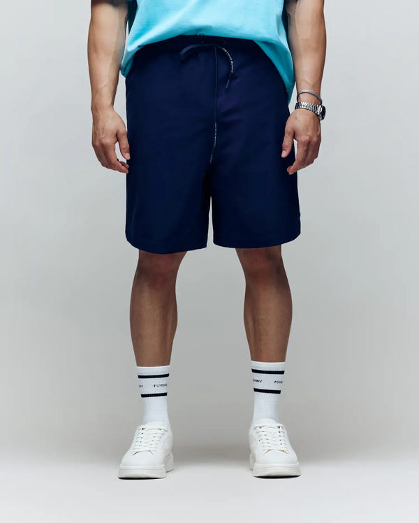 A person stands facing forward in a light blue shirt, Psycho Bunny MENS BIG AND TALL SAMOS HYDROCHROMIC SWIM TRUNK (B9W453F200), white sneakers, white socks with black stripes, and a wristwatch. The background is plain and neutral; face not visible.