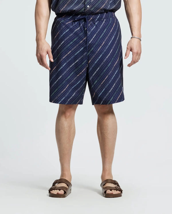 A person is wearing a Psycho Bunny patterned button-up shirt and the MENS BIG AND TALL RENMARK PB PRINTED SWIM TRUNK - B9W622F200 with navy quick-dry fabric featuring diagonal white and red stripes. They're paired with brown sandals against a plain light background.