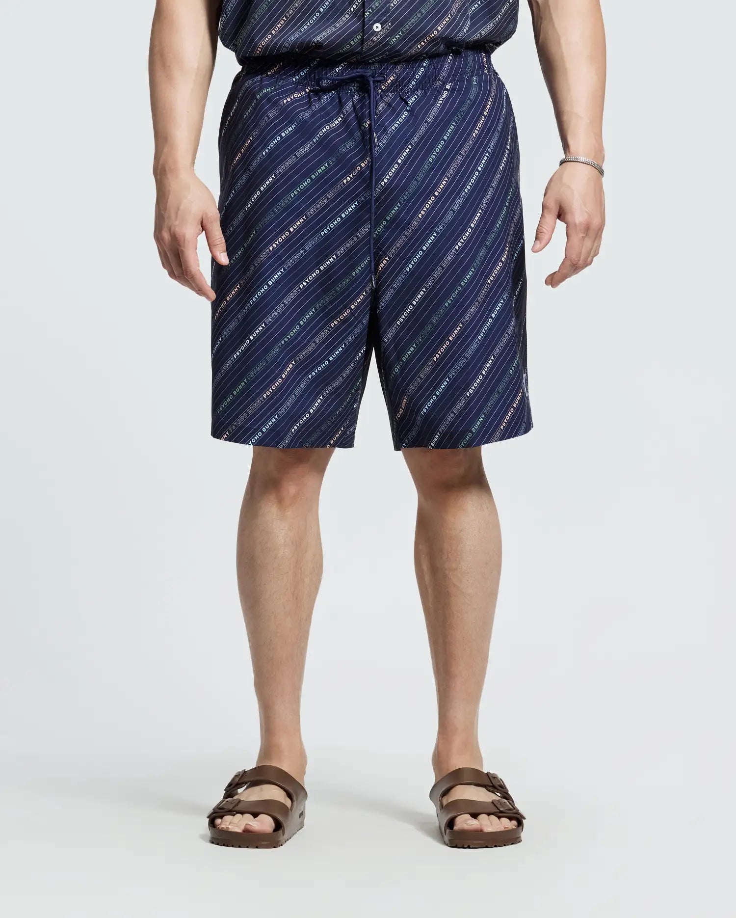 A person is wearing a Psycho Bunny patterned button-up shirt and the MENS BIG AND TALL RENMARK PB PRINTED SWIM TRUNK - B9W622F200 with navy quick-dry fabric featuring diagonal white and red stripes. They're paired with brown sandals against a plain light background.