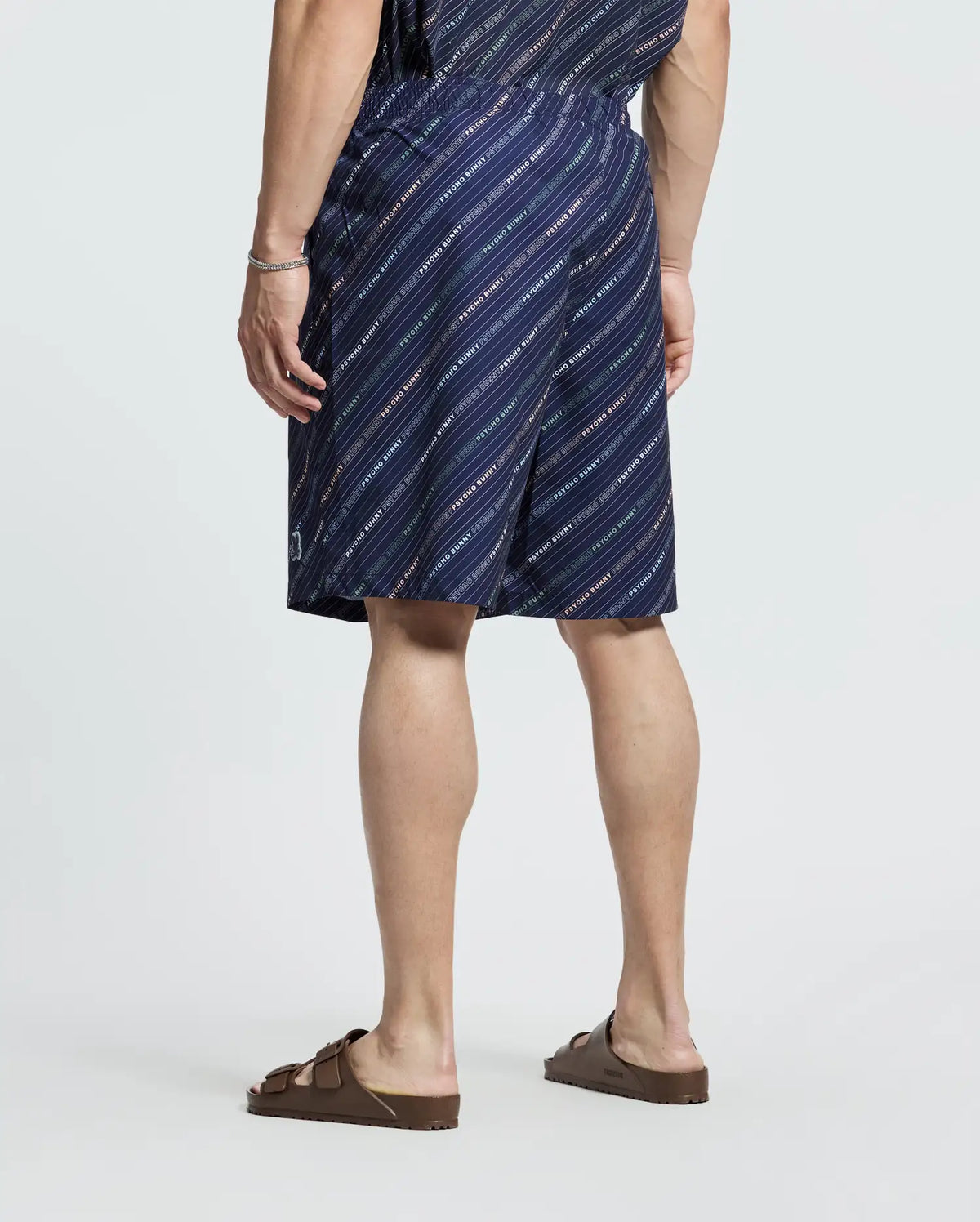 A person is seen from the back wearing the Psycho Bunny MENS BIG AND TALL RENMARK PB PRINTED SWIM TRUNK, with navy blue featuring diagonal white stripes, a logo-patterned top, and brown sandals against a plain background, embodying a casual style.