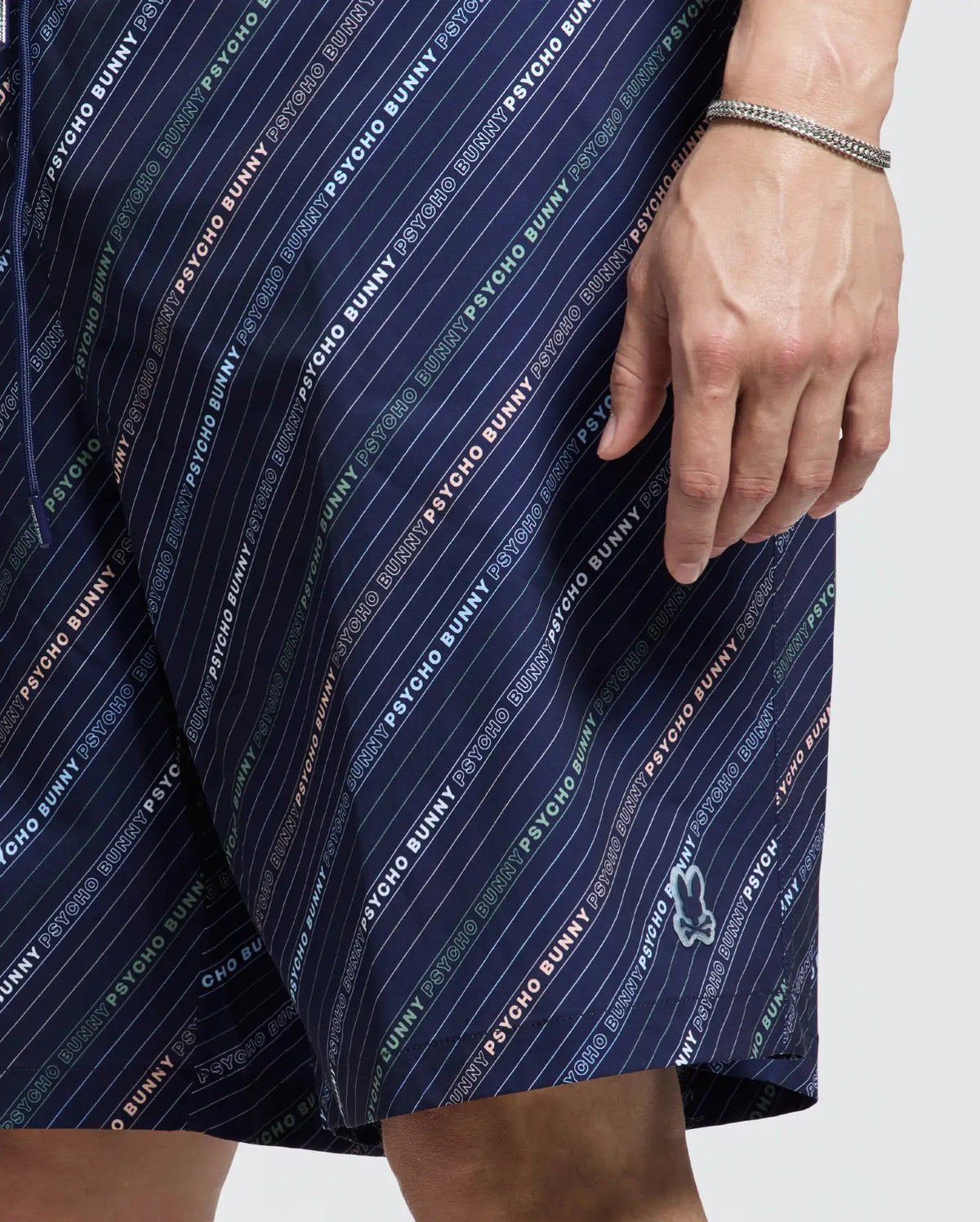 A close-up shows a person wearing Psycho Bunny's navy "MENS BIG AND TALL RENMARK PB PRINTED SWIM TRUNK," featuring diagonal stripes and a multi-color "PSYCHO BUNNY" logo print. A small bunny logo is near the hem, and their wrist is adorned with a bracelet.