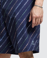 A close-up shows a person wearing Psycho Bunny's navy "MENS BIG AND TALL RENMARK PB PRINTED SWIM TRUNK," featuring diagonal stripes and a multi-color "PSYCHO BUNNY" logo print. A small bunny logo is near the hem, and their wrist is adorned with a bracelet.