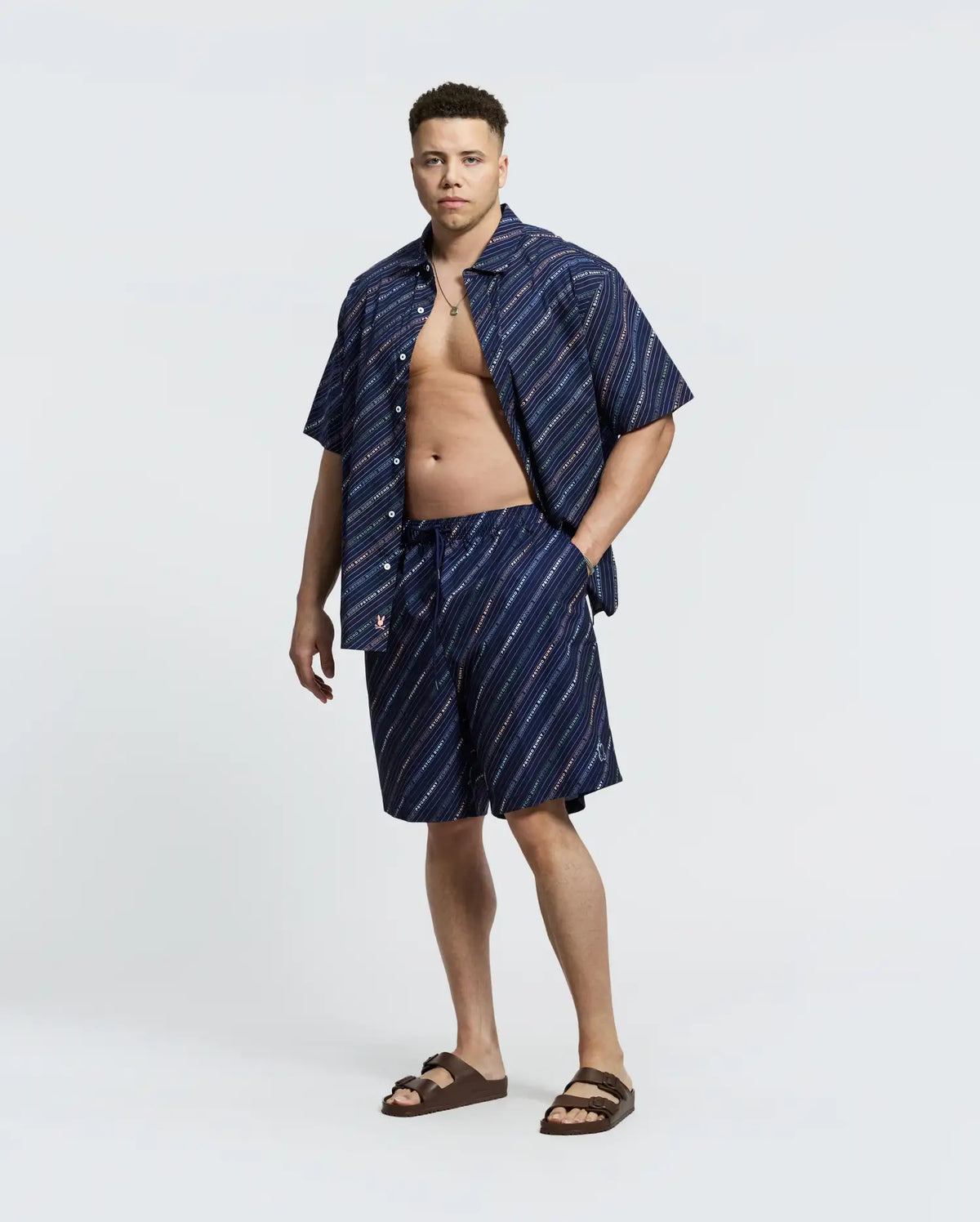 A man confidently stands against a plain background, wearing Psycho Bunny's blue striped shirt open to reveal his chest, paired with MENS BIG AND TALL RENMARK PB PRINTED SWIM TRUNK - B9W622F200. Brown sandals and a hand in his pocket complete his casual style for sunny adventures.
