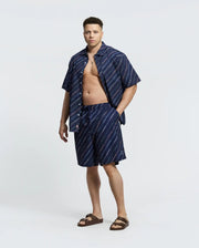 A man confidently stands against a plain background, wearing Psycho Bunny's blue striped shirt open to reveal his chest, paired with MENS BIG AND TALL RENMARK PB PRINTED SWIM TRUNK - B9W622F200. Brown sandals and a hand in his pocket complete his casual style for sunny adventures.