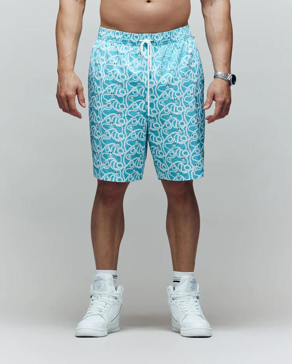 A man stands against a plain background wearing Psycho Bunny MENS BIG AND TALL CYRUS ALL OVER PRINT SWIM TRUNK - BB1000003, white high-top sneakers, and a watch. His upper body is not shown.