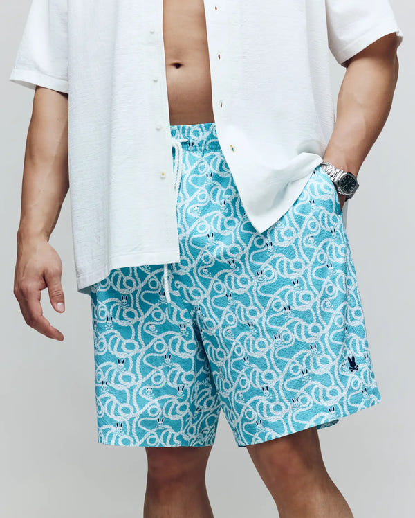 A person exudes coastal cool in Psycho Bunny’s MENS BIG AND TALL CYRUS ALL OVER PRINT SWIM TRUNK – BB1000003, featuring a light blue pattern with a white drawstring, paired with a crisp white short-sleeve button-up shirt and wristwatch, one hand in pocket.