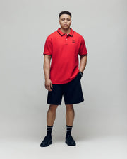 A man with a neutral expression models the Psycho Bunny MENS BIG & TALL JUDE PIQUE POLO - BT0100003 in red Pima cotton, paired with black shorts, striped black socks, and black shoes. An embroidered Mischief Bunny lends a playful accent.