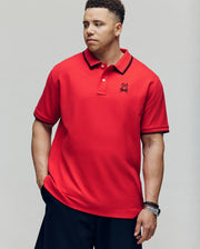 A man in the Psycho Bunny MENS BIG & TALL JUDE PIQUE POLO (BT0100003), made of soft Pima cotton with black trim and an embroidered Mischief Bunny logo, stands with one hand in his pocket against a plain background.