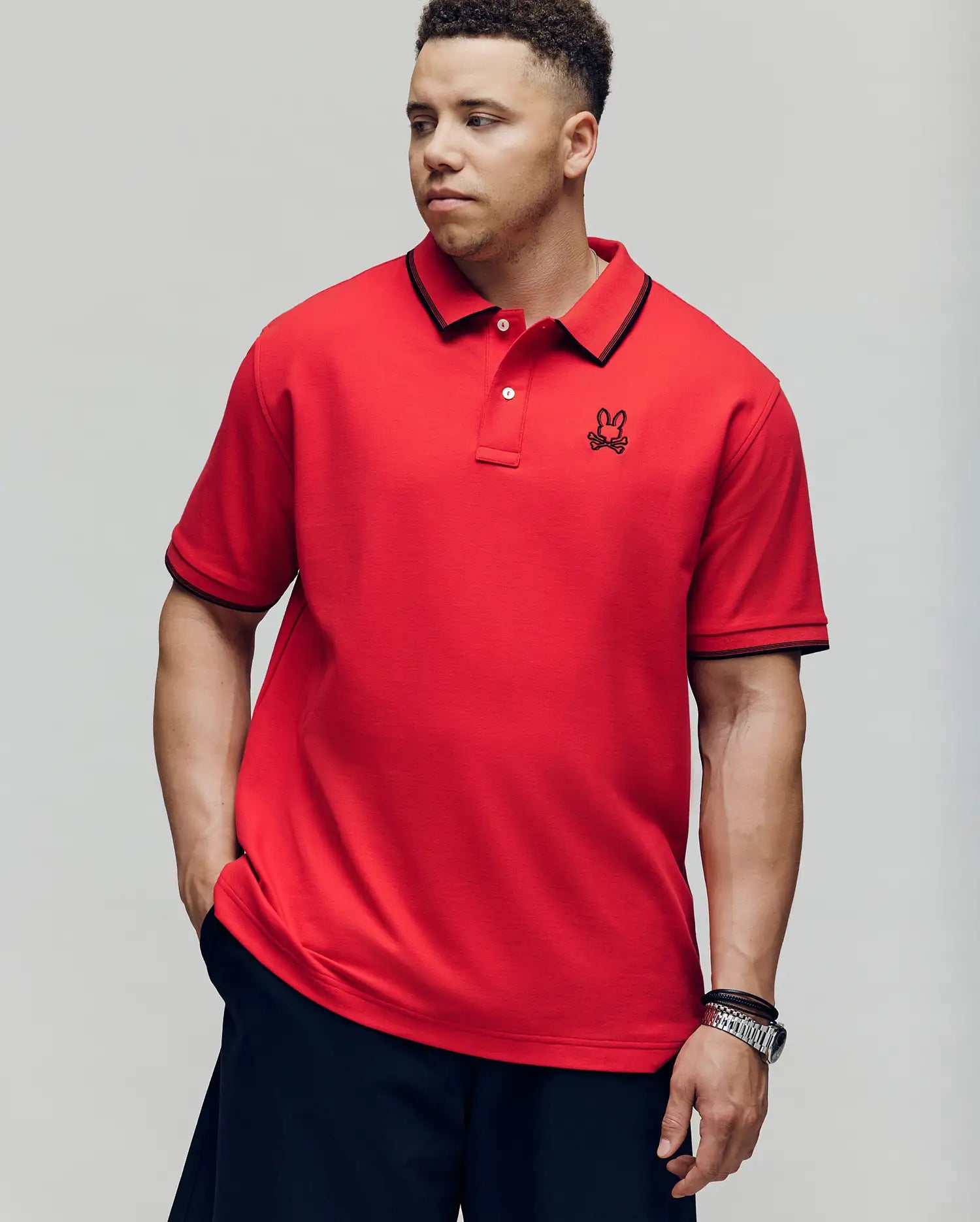 A man in the Psycho Bunny MENS BIG & TALL JUDE PIQUE POLO (BT0100003), made of soft Pima cotton with black trim and an embroidered Mischief Bunny logo, stands with one hand in his pocket against a plain background.
