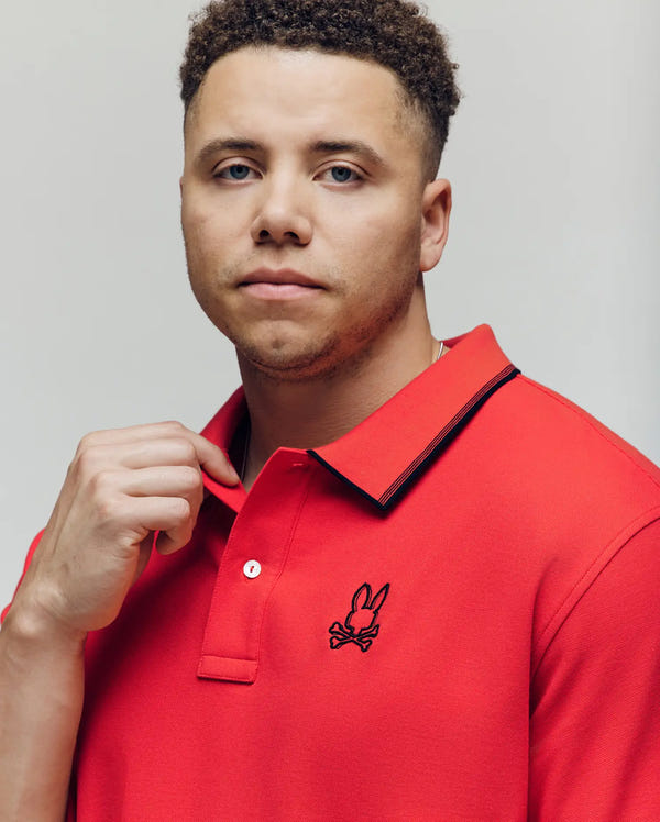 A man with short curly hair wears the Psycho Bunny MENS BIG & TALL JUDE PIQUE POLO (BT0100003) with an embroidered Mischief Bunny logo, standing against a light background and looking calmly at the camera.