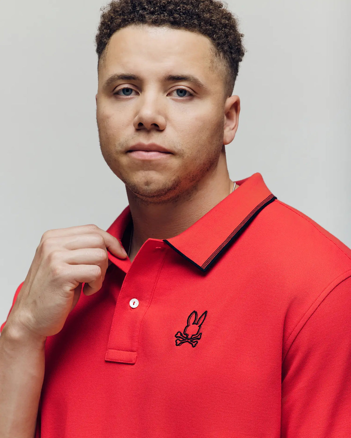 A man with short curly hair wears the Psycho Bunny MENS BIG & TALL JUDE PIQUE POLO (BT0100003) with an embroidered Mischief Bunny logo, standing against a light background and looking calmly at the camera.