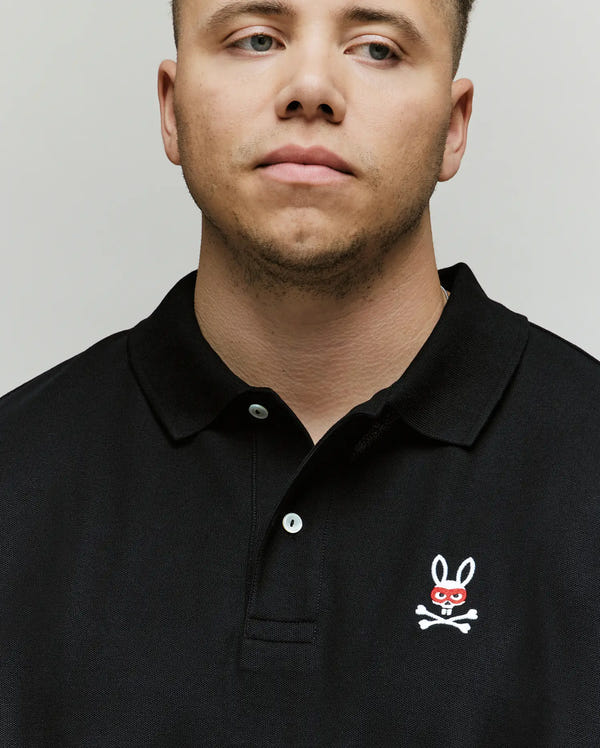 A man in the Psycho Bunny MENS BIG & TALL MISCHIEF PIQUE POLO (BT0100006) with an embroidered bunny face, red eyeglasses, and crossbones on the chest, poses slightly to the side against a plain background.