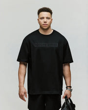 A man with short curly hair wears a relaxed-fit black Psycho Bunny MENS BIG AND TALL ZAYN OVERSIZED GRAPHIC TEE (BT0200009) and matching shorts, holding a black bag and standing against a plain light background.