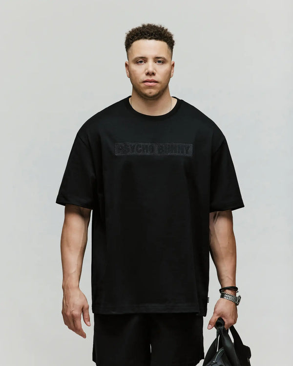 A man with short curly hair wears a relaxed-fit black Psycho Bunny MENS BIG AND TALL ZAYN OVERSIZED GRAPHIC TEE (BT0200009) and matching shorts, holding a black bag and standing against a plain light background.