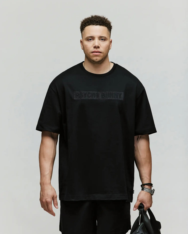 A man with short curly hair wears a relaxed-fit black Psycho Bunny MENS BIG AND TALL ZAYN OVERSIZED GRAPHIC TEE (BT0200009) and matching shorts, holding a black bag and standing against a plain light background.