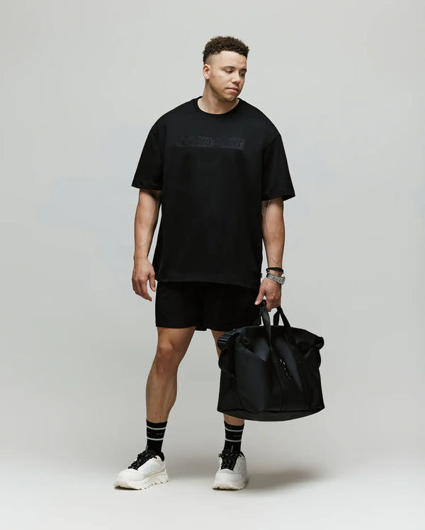 A man stands against a plain background wearing a Psycho Bunny MENS BIG AND TALL ZAYN OVERSIZED GRAPHIC TEE (BT0200009), black shorts, black socks, and white sneakers. He holds a large black bag and looks downward with a relaxed expression.