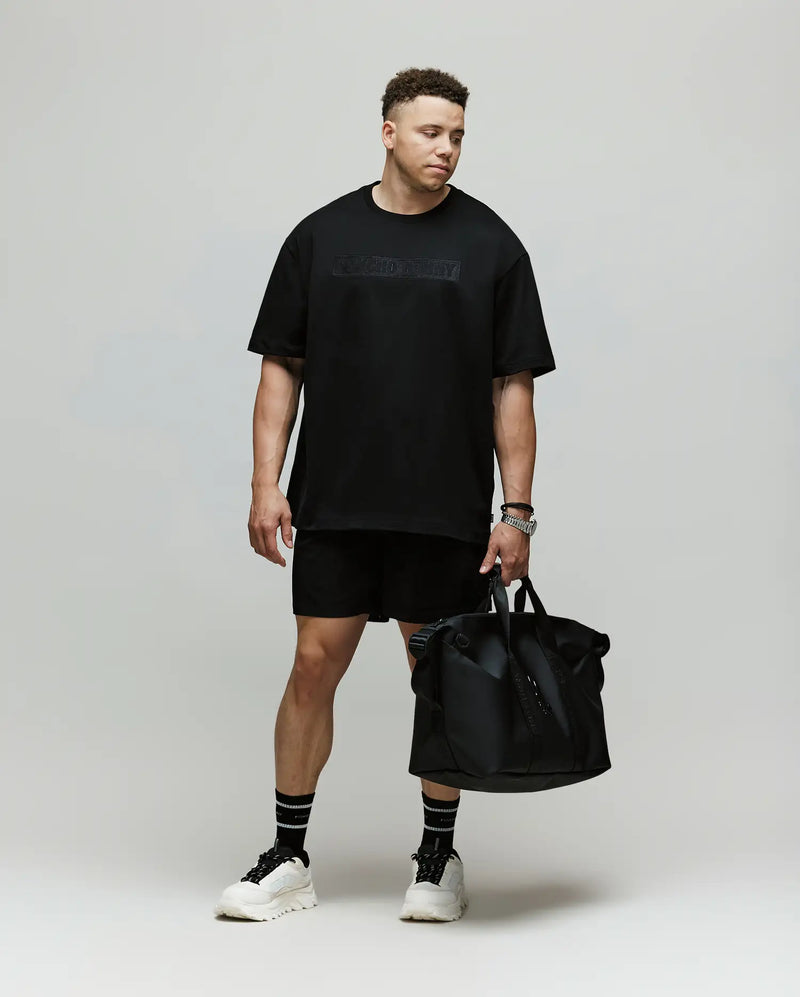 A man stands against a plain background wearing a Psycho Bunny MENS BIG AND TALL ZAYN OVERSIZED GRAPHIC TEE (BT0200009), black shorts, black socks, and white sneakers. He holds a large black bag and looks downward with a relaxed expression.