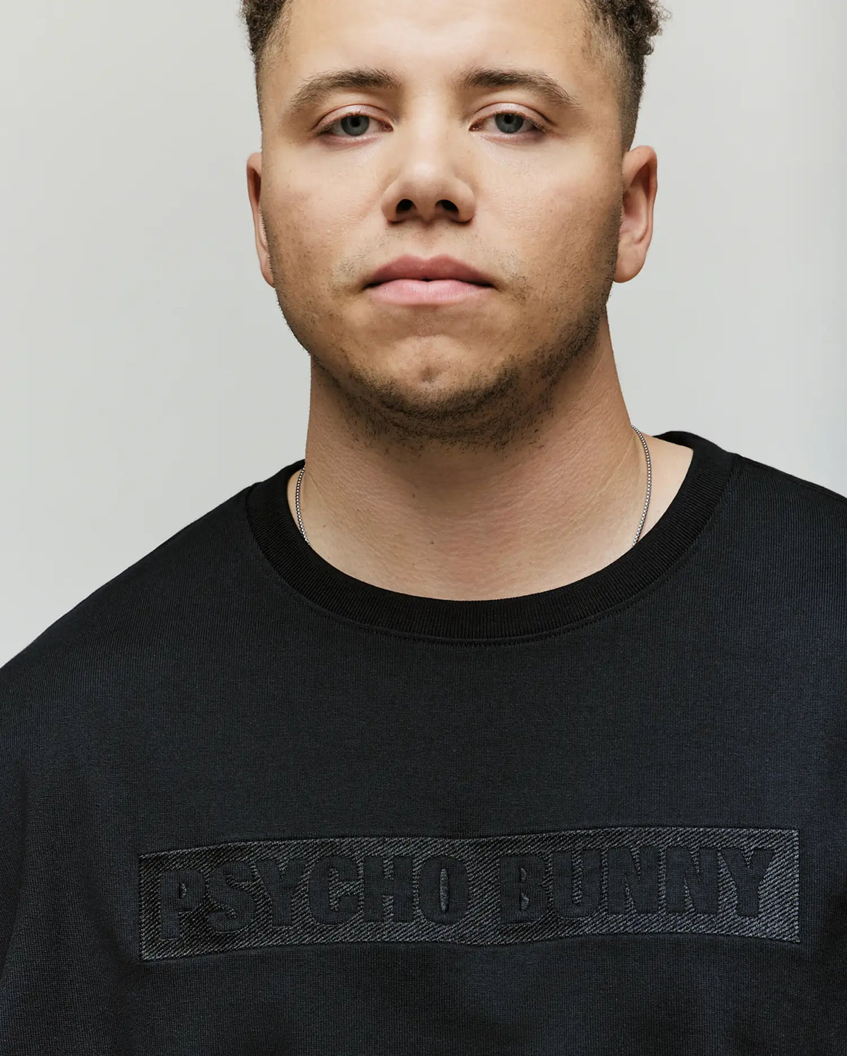 A person wears the Psycho Bunny Mens Big and Tall Zayn Oversized Graphic Tee (BT0200009) in black, featuring front embroidery and a relaxed fit, while looking at the camera against a plain light background.