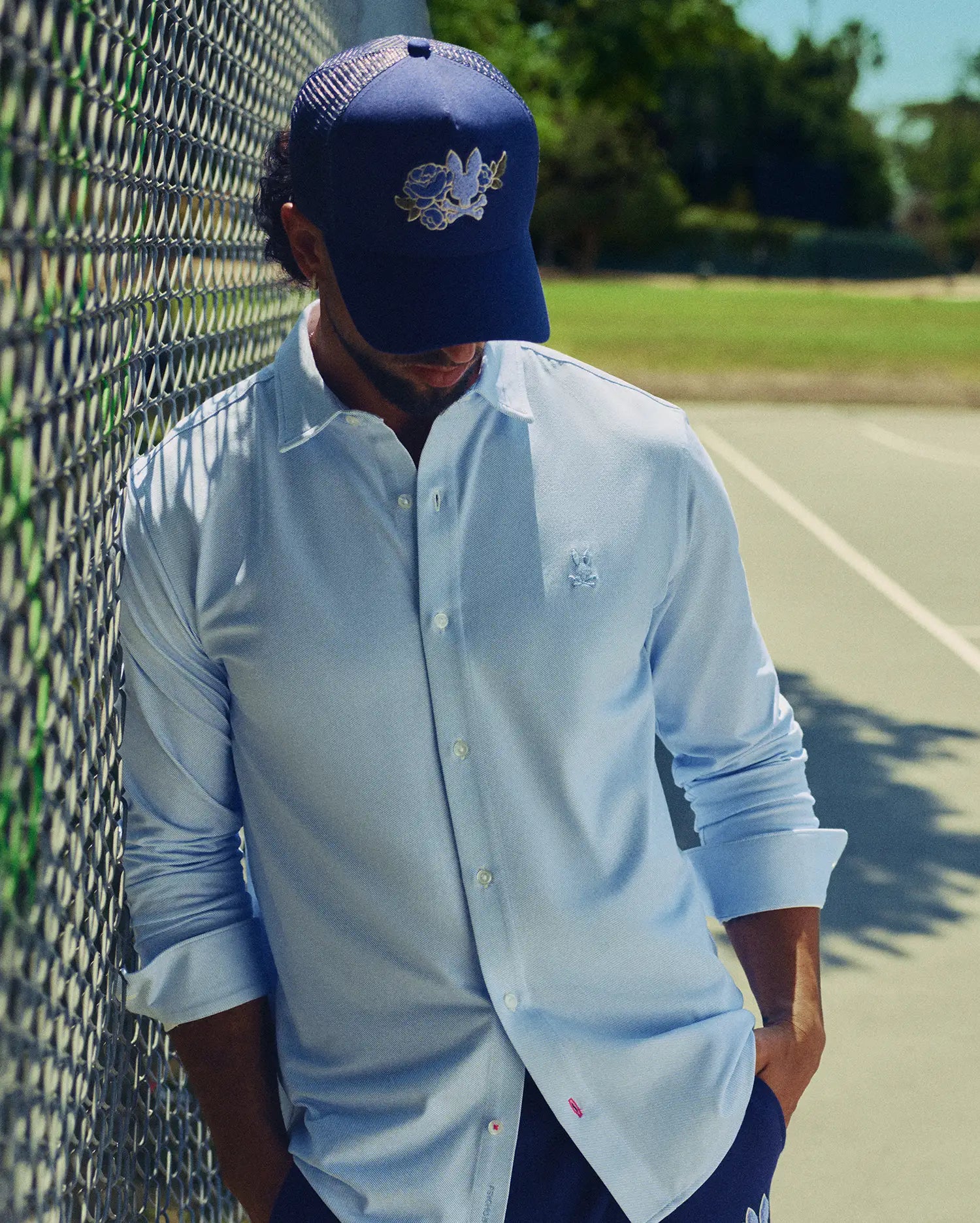 A man in a light blue button-down and the Psycho Bunny MENS AUGUSTE TRUCKER CAP (MO1500050) stands by a chain-link fence on a sunny day, with a tennis court and trees behind him.