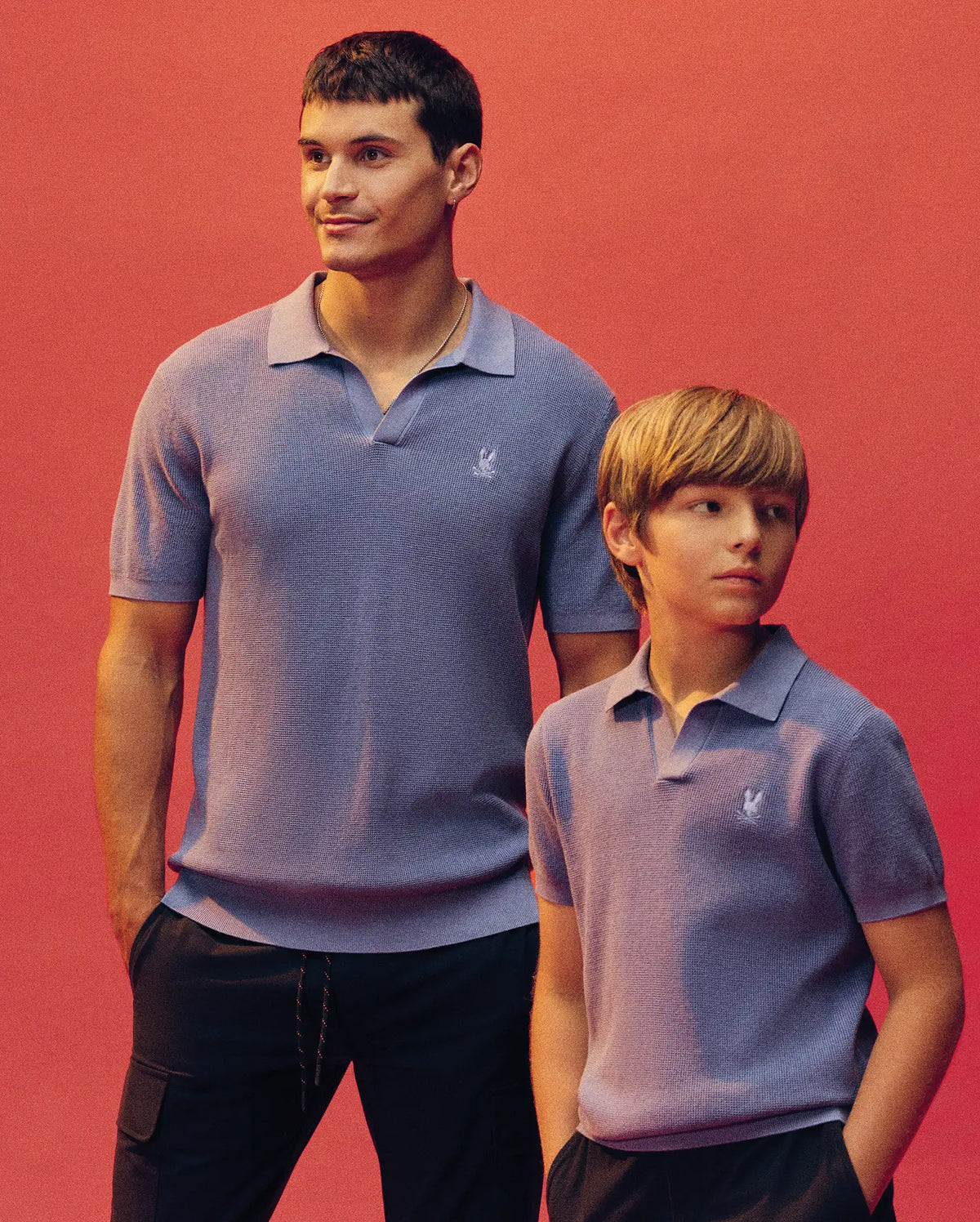 An adult and child, both in matching Psycho Bunny KIDS LEETON KNIT POLO SHIRTS (B0E562F200) in light purple, stand against a coral pink background. The adult smiles and looks up as the child glances to the side.
