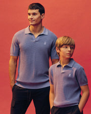 An adult and child, both in matching Psycho Bunny KIDS LEETON KNIT POLO SHIRTS (B0E562F200) in light purple, stand against a coral pink background. The adult smiles and looks up as the child glances to the side.