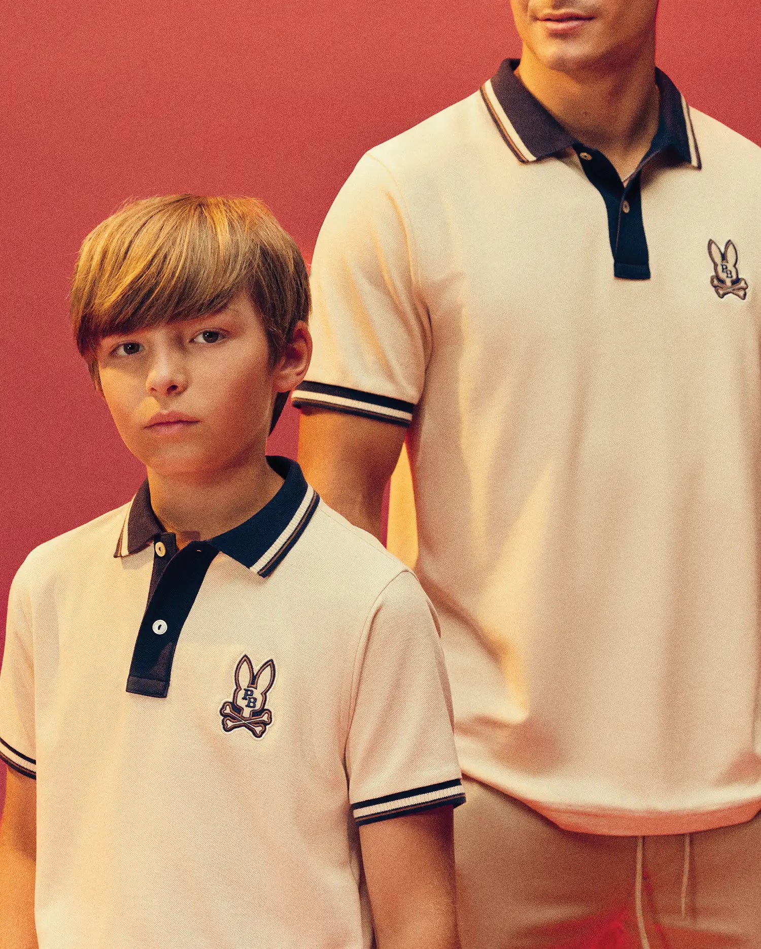 A young boy and an adult man wear matching Psycho Bunny KIDS WADE PIQUE POLO SHIRTS (KT0100032) with black collars, colorblock tipping, and a bunny skull chest logo, set against a warm reddish background.