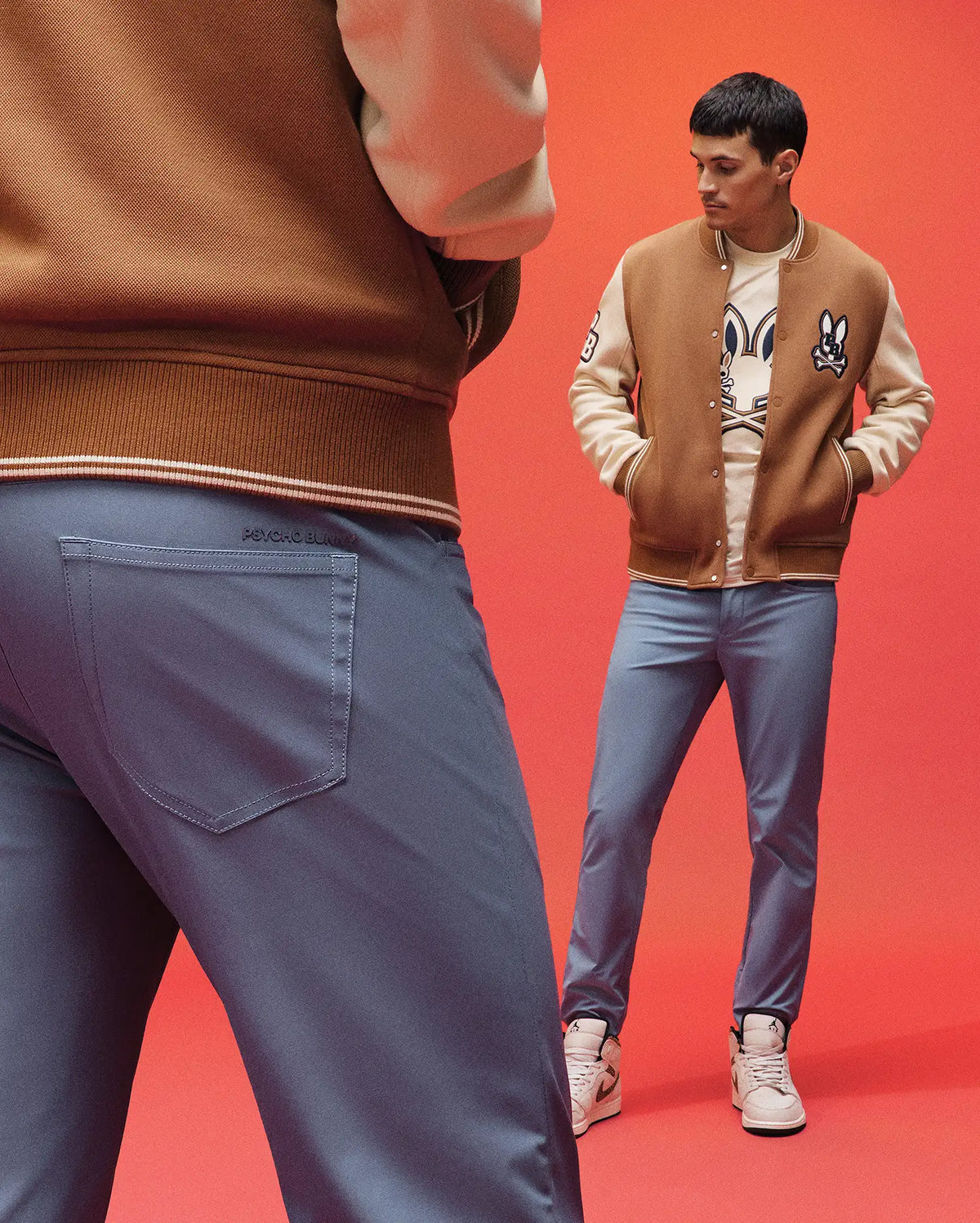 Two men in brown varsity jackets, sneakers, and Psycho Bunny MENS MAVERICK 10K STEP PANT - B6P788D200, stand against a solid red background—one faces away in the foreground as the other looks at him from afar.