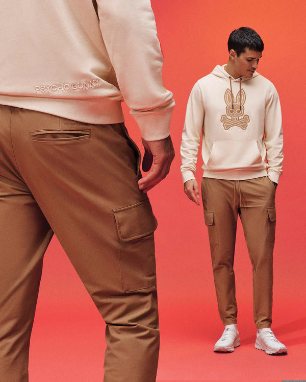 Two men model Psycho Bunny's MENS LLOYD CARGO PANT (B6P159B200) in beige with hoodies—one with a bunny logo—against a red backdrop. One faces front, the other turns his back; both wear white sneakers.