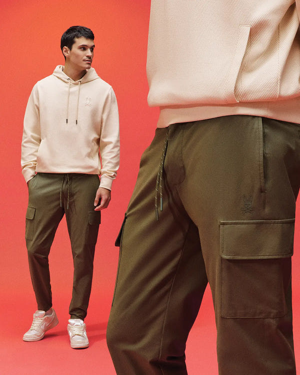 A man in a cream hoodie and white sneakers stands against a bright red background, wearing Psycho Bunny’s MENS LLOYD CARGO PANT - B6P159B200; the foreground highlights the pants’ pockets and drawstrings.