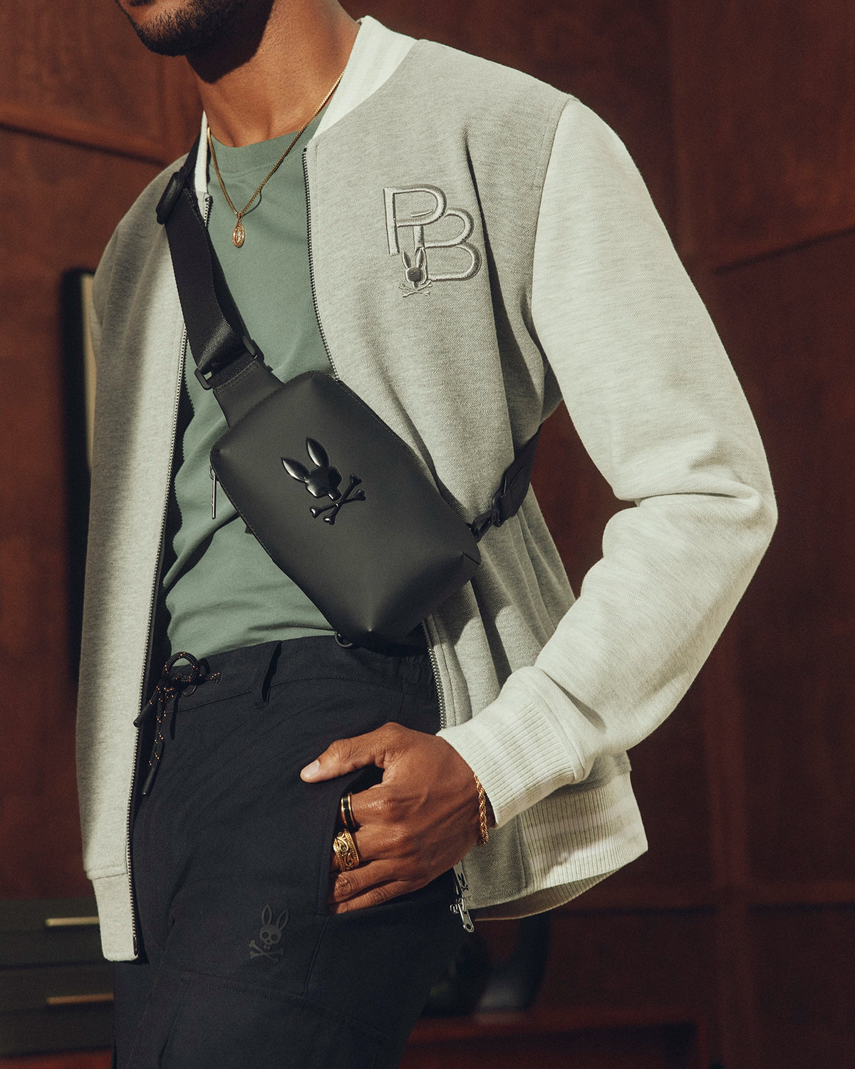 A person is wearing a light gray "PB"-embroidered bomber jacket, green shirt, and black pants, accessorized with gold jewelry. They carry Psycho Bunny's Men's Rubberized Crossbody Bag - B6A635F200 against a warm-toned interior backdrop.