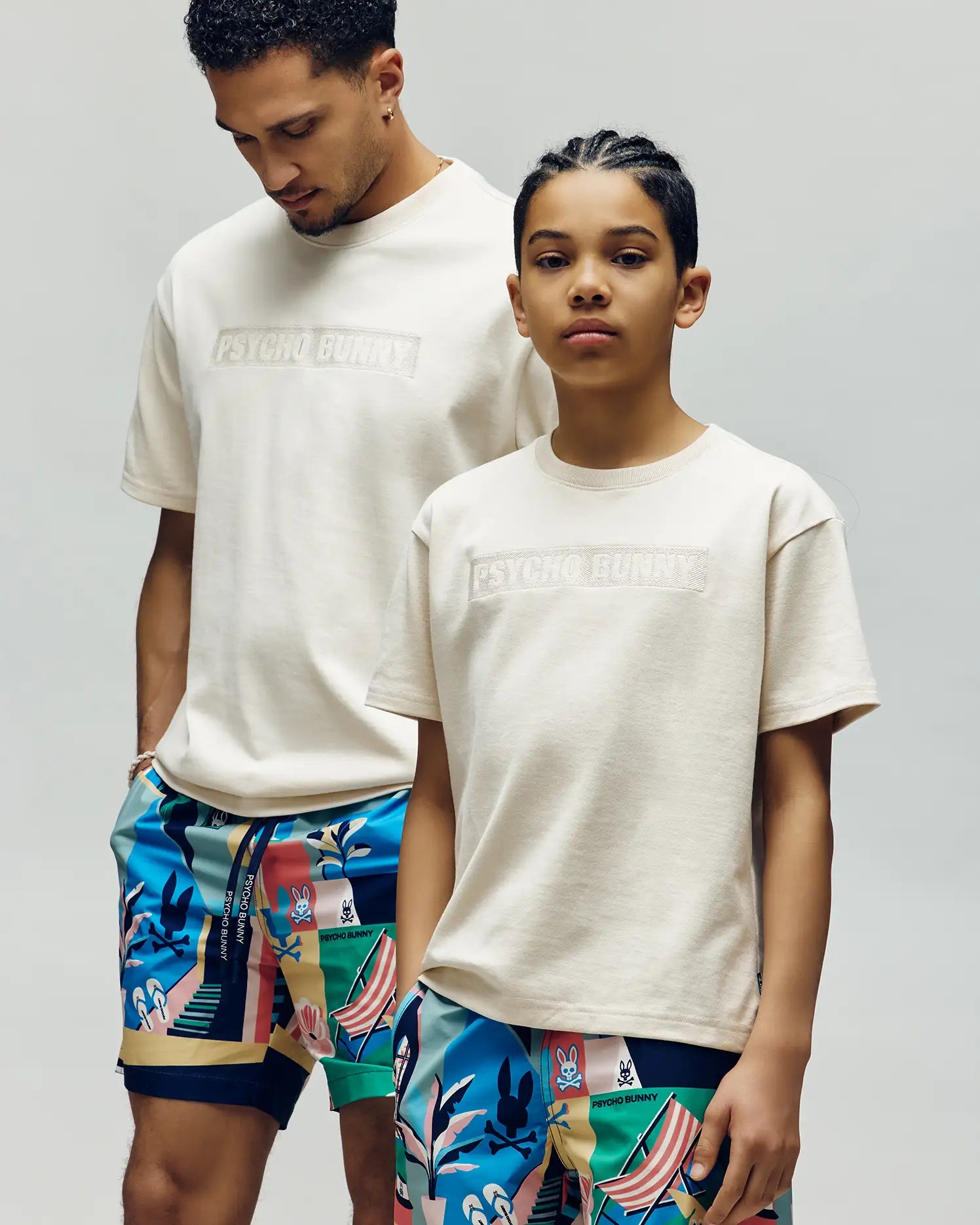 Two kids wear Psycho Bunny KIDS ZAYN OVERSIZED GRAPHIC TEE in off-white, styled with colorful shorts. Against a plain background, one looks down while the other faces forward with a neutral expression.