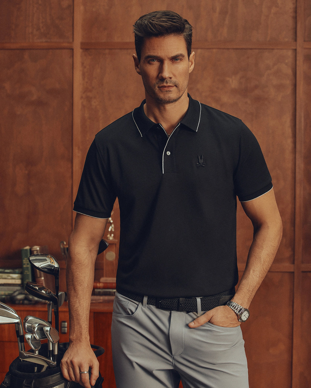 Indoors, a man in a Psycho Bunny MENS BRYCE PERFORMANCE PIQUE POLO - B6K486F200 stands in gray pants, resting one hand on a golf club. He has short dark hair and is clean-shaven. Books and decor can be seen on a wooden surface in the background.