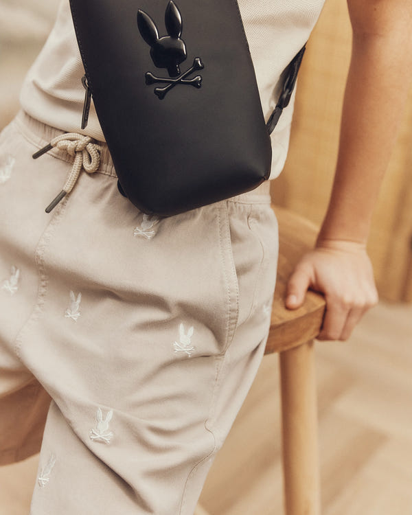 A person in beige KIDS ALL OVER WILLIS STRETCH TENCEL SHORT - B0R559F200 with a bunny print, a cream top, and a black crossbody bag with a bunny and crossbones emblem leans against a wooden chair in warm lighting, exuding casual charm in the regular-fit Psycho Bunny ensemble.