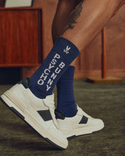 A close-up shows someone sporting Psycho Bunny's MENS 3 PACK DRESS SOCK - B6F504F200 in blue, paired with white sneakers featuring black accents. A tattoo adds a unique touch to the look against a wooden cabinet and brown wall backdrop.
