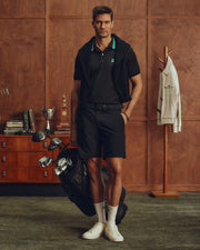 A man stands in a wooden-walled room, wearing a Psycho Bunny MENS FORREST SPORT PIQUE POLO - B6K660F200, shorts, white sneakers, and high socks. He carries a golf bag with clubs. To his left are trophies on furniture and a coat on a wooden rack.