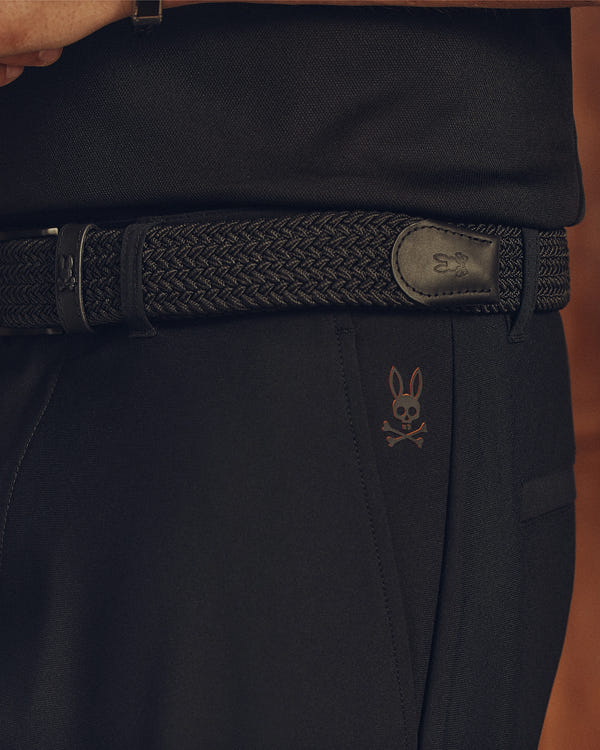 A close-up shows a person in durable black pants with a subtle bunny and crossbones logo, accessorized with a Psycho Bunny MENS STRETCH BRAIDED BELT (B6A890F200). The belt's buckle features the same logo, perfectly complementing the tucked-in black shirt.