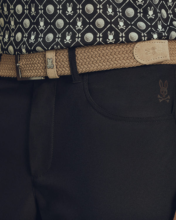 A stylish individual wears dark pants with a small skull and bunny logo, accessorized by a beige Psycho Bunny MENS STRETCH BRAIDED BELT (B6A890F200) featuring a matching emblem on the buckle. Their shirt's pattern of skulls and bunny ears completes the look seamlessly.