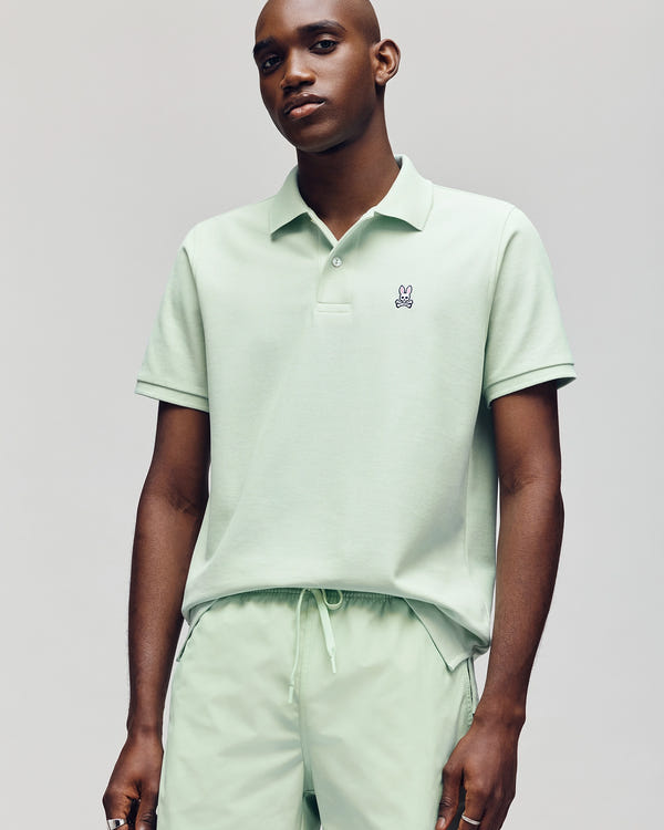 A man stands against a plain background in a Psycho Bunny Men's Classic Pique Polo, made from soft Pima Cotton with an embroidered bunny logo, pairing it with matching light green shorts and maintaining a neutral expression.