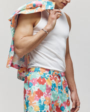 A person in Psycho Bunny's vibrant MENS LEMMON ALL OVER PRINT SWIM TRUNK, featuring a colorful floral pattern, holds a matching jacket. The ensemble, with shades of red, orange, pink, and blue against a plain backdrop, effortlessly combines comfort and style.