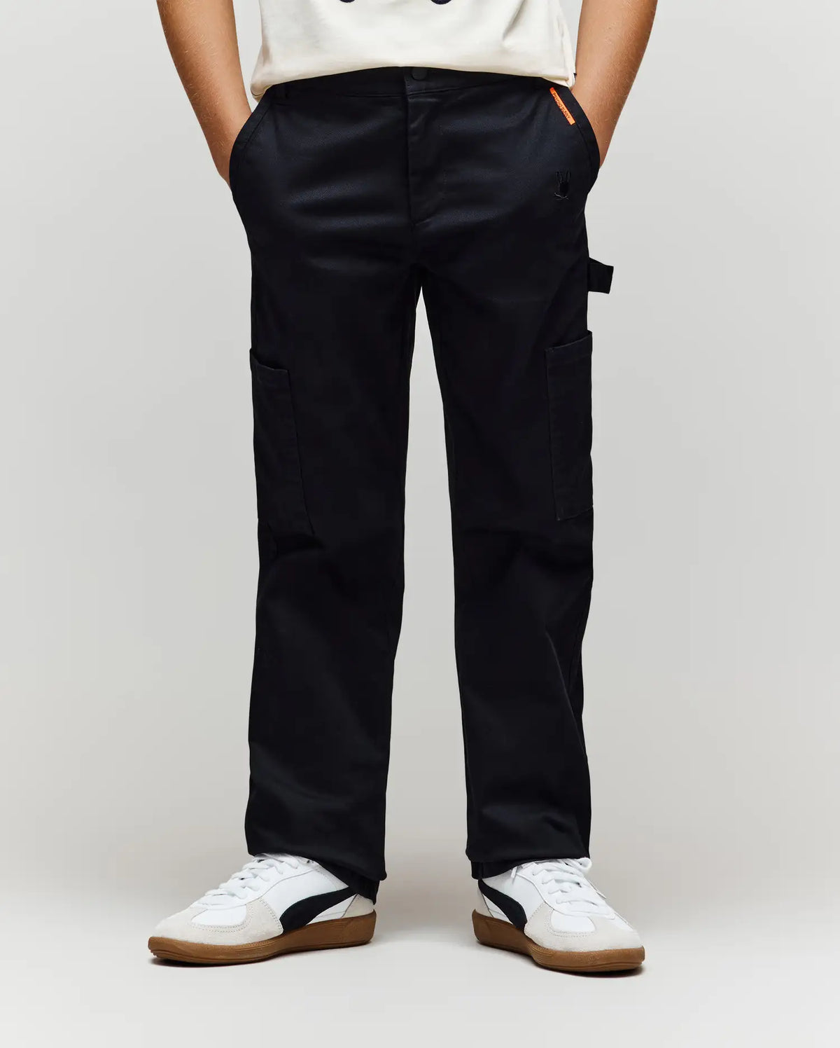 A person wearing Psycho Bunny KIDS RUSSELL CARPENTER PANT - KB0600001 in black, a white t-shirt, and white sneakers with black and brown details stands with hands in their pockets against a plain background.