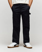 A person wearing Psycho Bunny KIDS RUSSELL CARPENTER PANT - KB0600001 in black, a white t-shirt, and white sneakers with black and brown details stands with hands in their pockets against a plain background.