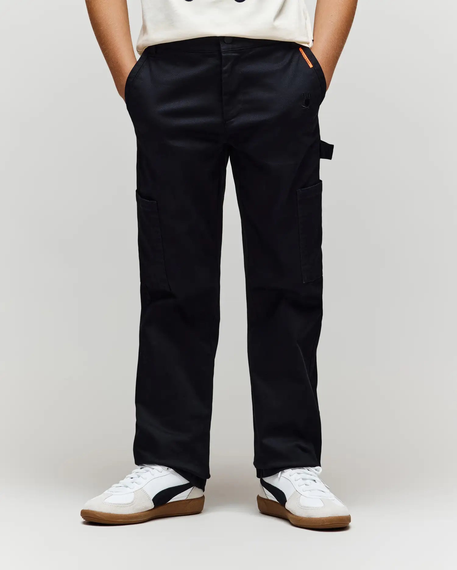 A person wearing Psycho Bunny KIDS RUSSELL CARPENTER PANT - KB0600001 in black, a white t-shirt, and white sneakers with black and brown details stands with hands in their pockets against a plain background.