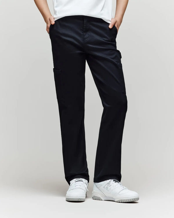 A person models the Psycho Bunny KIDS RUSSELL CARPENTER PANT (KB0600001) in black with cargo pockets and white sneakers, hands in pockets, against a simple light backdrop, showing only the lower half.