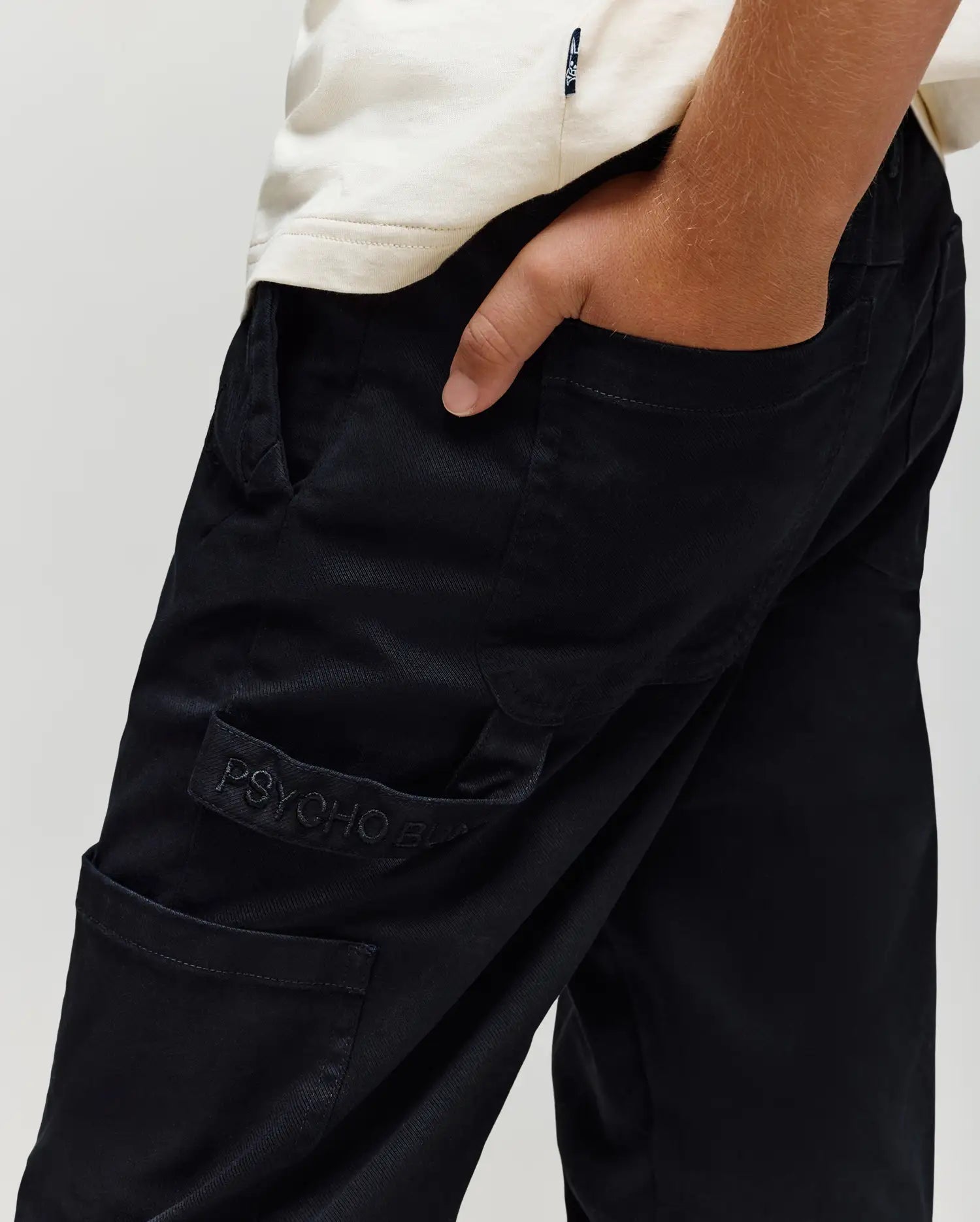 A person wears KIDS RUSSELL CARPENTER PANT - KB0600001 by Psycho Bunny in black, featuring a visible "PSYCHO" logo on the cargo pocket, one hand in the pocket, and paired with a white shirt.