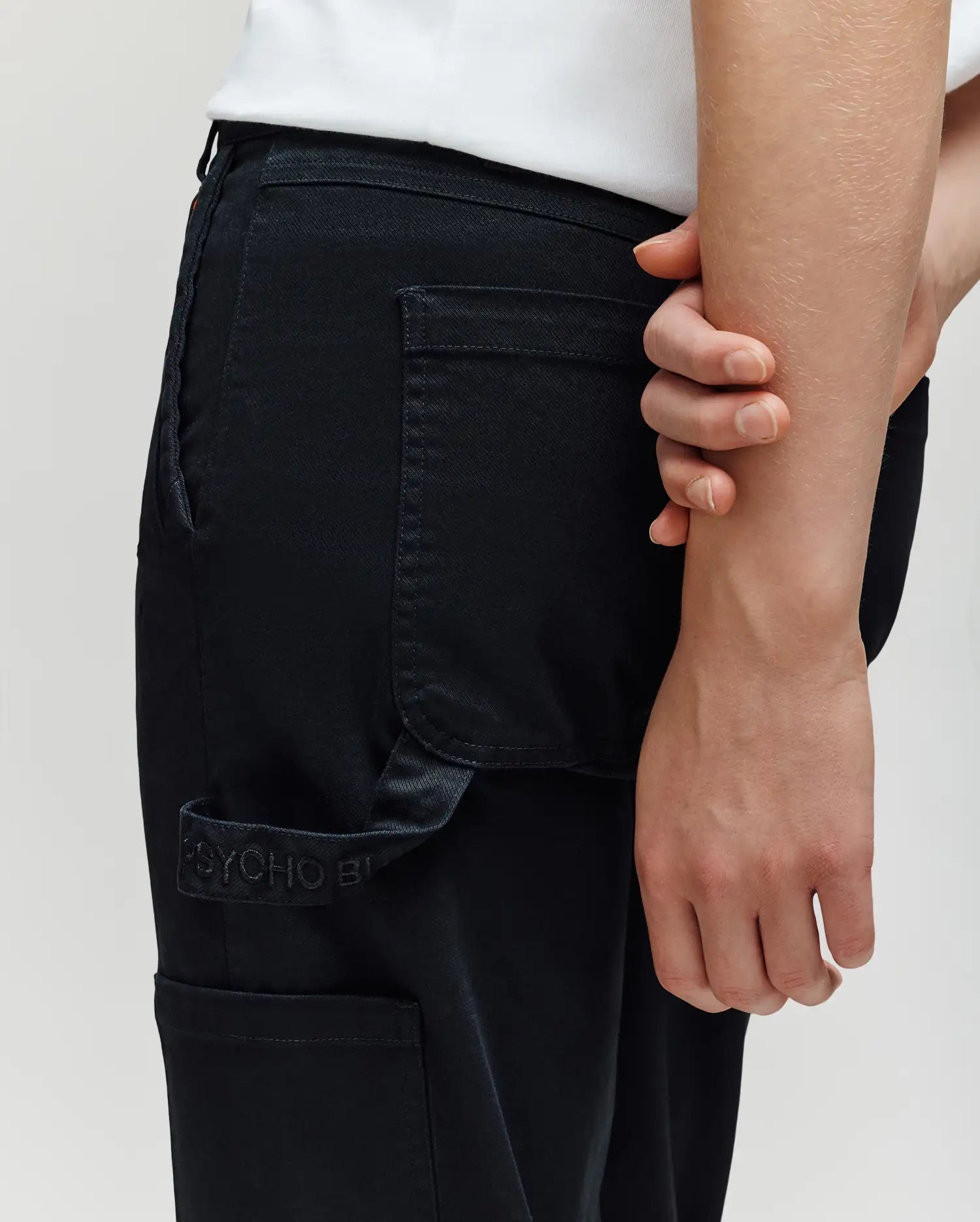 A person wears Psycho Bunny KIDS RUSSELL CARPENTER PANT (KB0600001) in black with a regular fit and visible back pocket, paired with a white shirt, standing with one arm bent behind their back and holding the opposite wrist.